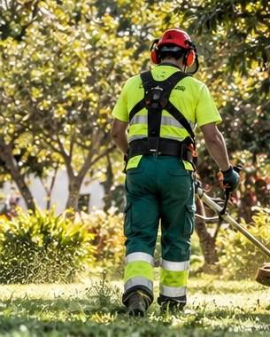 Operario con casco, protección auditiva y ropa de alta visibilidad desbrozando el césped de un parque arbolado.
