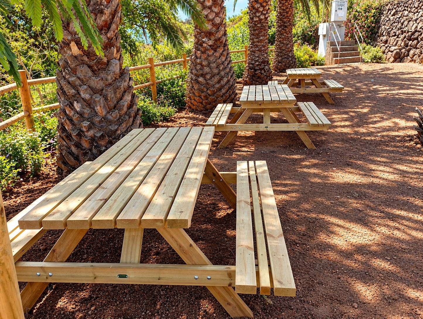 Mesas de picnic de madera nuevas bajo unas palmeras, en un área recreativa.